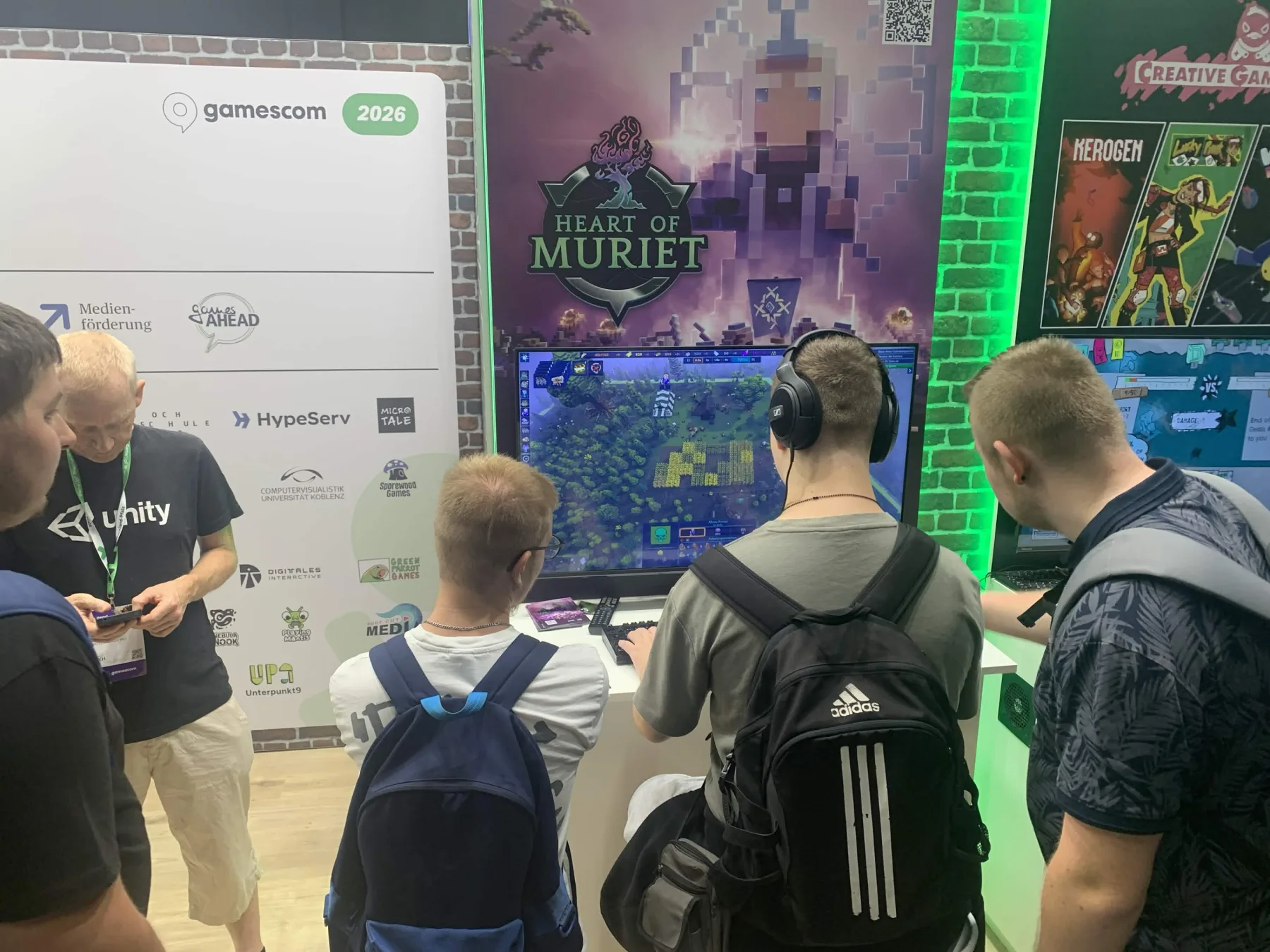 Visitors playing the Heart of Muriet demo at the gamescom 2026 booth, with the game banner and sponsor wall behind them